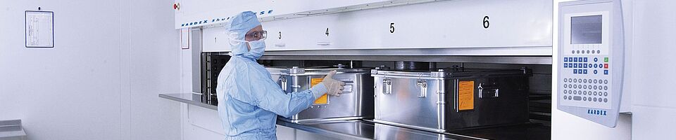 Man in full laboratory clothing at a cleanroom lift system with several metal containers.
