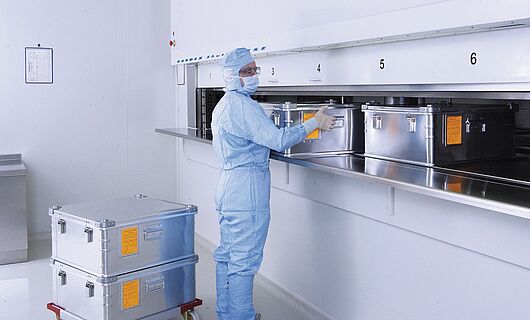 Mann in voller Laborkleidung an einem Reinraum-Liftsystem mit mehreren Metallkisten.