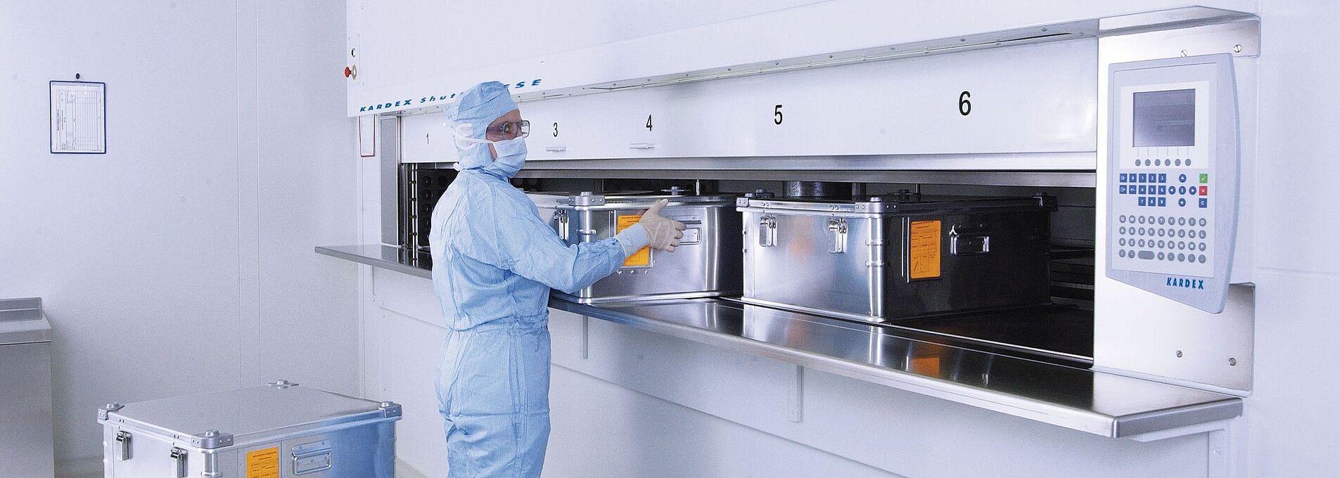 Man in full laboratory clothing at a cleanroom lift system with several metal containers.