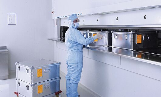 Man in full laboratory clothing at a cleanroom lift system with several metal containers.
