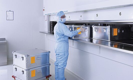 Mann in voller Laborkleidung an einem Reinraum-Liftsystem mit mehreren Metallkisten.