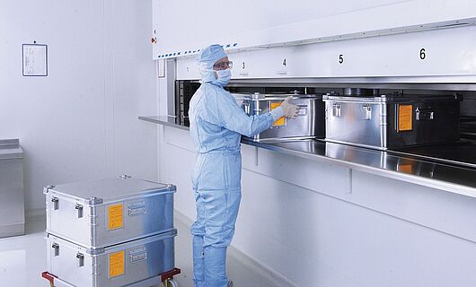 Man in full laboratory clothing at a cleanroom lift system with several metal containers.