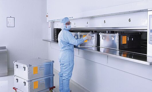 Mann in voller Laborkleidung an einem Reinraum-Liftsystem mit mehreren Metallkisten.
