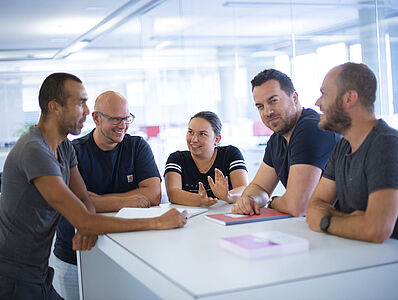 Five people are standing together at a standing table, engaged in a discussion.