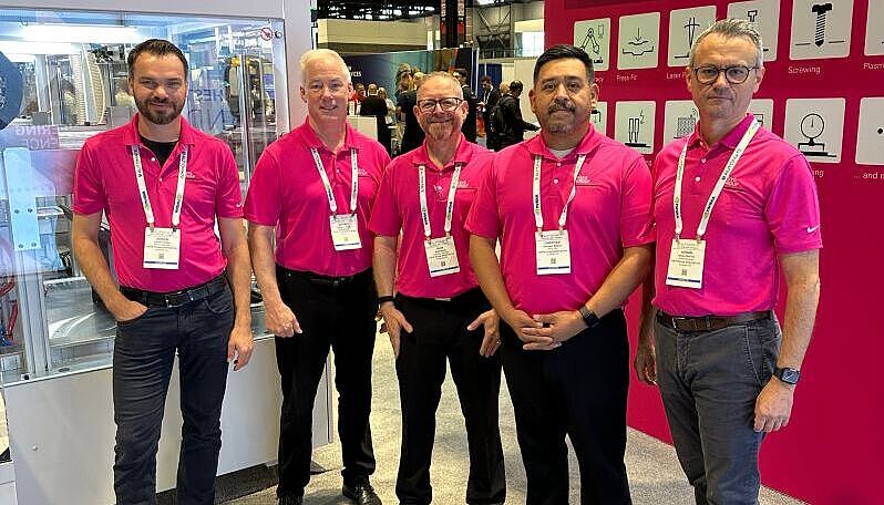 Five men standing side by side in magenta polo shirts at a trade show booth.