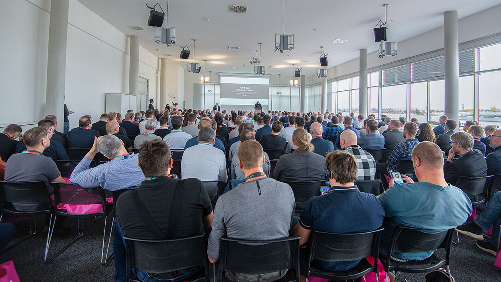 A rear view of a large conference room with many people seated. One person is giving a presentation on a stage.