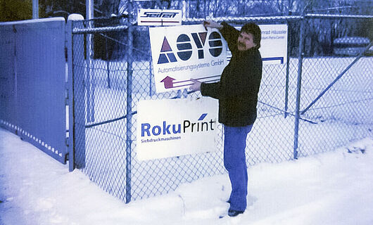 Ein Mann steht im Schnee und befestigt ein Schild an einen Zaun. Jahr 1996