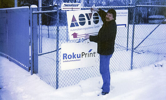 A man stands in the snow, attaching a sign to a fence. Year 1996