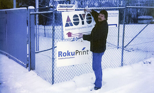 Ein Mann steht im Schnee und befestigt ein Schild an einen Zaun. Jahr 1996