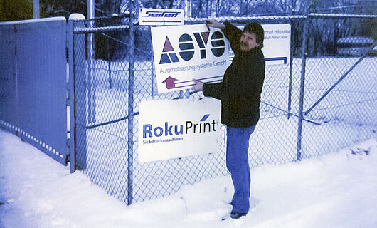 Ein Mann steht im Schnee und befestigt ein Schild an einen Zaun. Jahr 1996