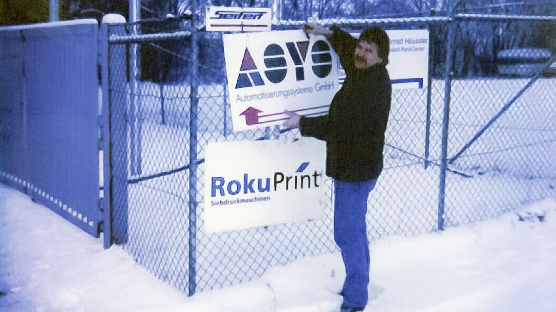 Ein Mann steht im Schnee und befestigt ein Schild an einen Zaun. Jahr 1996